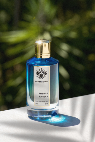 Bottiglia di profumo Mancera French Riviera da 120 ml, fotografata al sole con riflessi dorati, su sfondo verde ispirato alla natura mediterranea.